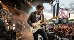 A British indie rock musician performing live on stage at a UK festival playing a classic Olympic white Fender Stratocaster.